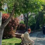 Elderly man in a beige suit sitting quietly on a bench under leafy trees in a peaceful park
